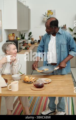 Kaukasische Frau, die eine Kaffeetasse hält, während ein afroamerikanischer Mann mit einem Frühstücksteller steht, der beide lächelt und sich in einem Gespräch befindet Stockfoto