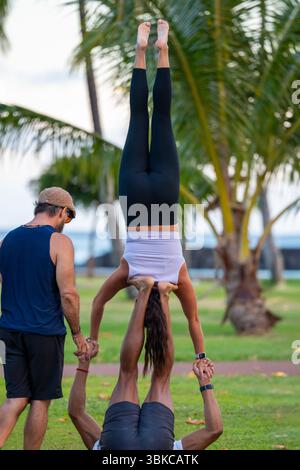 Eine Frau führt einen unterstützten Akro-Yoga-Handstand auf, der von einem Partner gehalten wird, während ein Spotter vor einem tropischen Park hilft. Ein Moment des Gleichgewichts Stockfoto