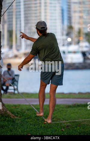 Ein Mann balanciert barfuß auf einer Slackline zwischen Bäumen in der Nähe eines Yachthafens. Das Bild erfasst Fokus, Koordination und Erholung im Freien Stockfoto