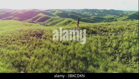 Aus der Vogelperspektive eine junge Frau, die ihr Trekkingabenteuer genießt, während sie bei einem atemberaubenden Sonnenuntergang über die üppigen, grasbewachsenen Hügel der Insel Sumba, Indonesien, spaziert Stockfoto