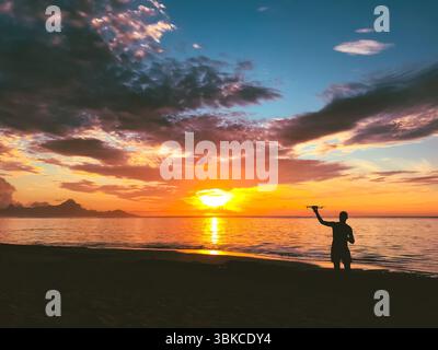 Ein Mann, der den Sonnenuntergang mit Drohne schießt. Die Silhouette eines Fahrers, der Quadracopter in der Hand hält und den hellorangefarbenen Sonnenuntergang über dem Meer und den bewölkten Himmel beobachtet. Wunderschöne Naturlandschaft. Beliebtes Touristenziel Stockfoto