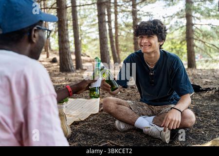 Zwei fröhliche, vielfältige junge männliche Freunde trinken mit Bierflaschen und feiern ihre Freundschaft während eines Campingausflugs in der Natur Stockfoto