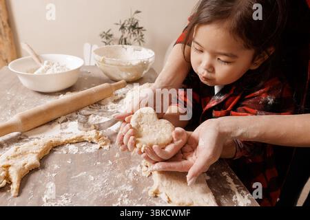 Kind und Mutter backen zusammen, halten herzförmigen Teig und symbolisieren Familienliebe, Sorgfalt und Freude in einer gemütlichen Küche. Stockfoto