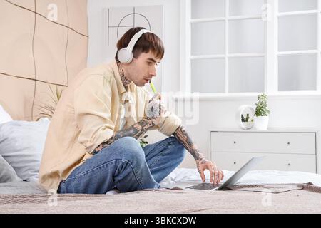 Junger gutaussehender Mann in Kopfhörern mit elektronischer Wegwerfzigarette, der Laptop auf dem Bett zu Hause benutzt Stockfoto