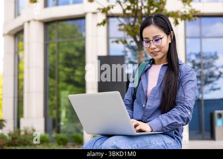Eine junge Asiatin benutzt draußen einen Laptop, mit einem Gebäude und Grün im Hintergrund. Stockfoto