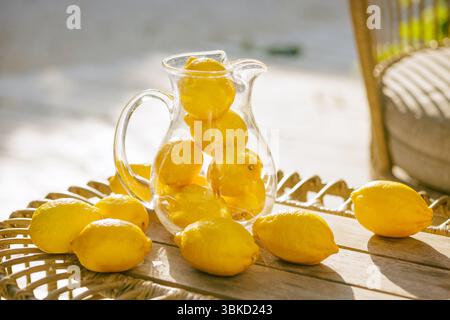 Glaskanne gefüllt mit frischen Zitronen auf einem Korbtisch, lebhaftes Sommerzitrusarrangement in hellem natürlichem Sonnenlicht. Stockfoto