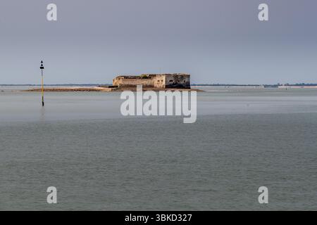 Fort Énet liegt vor Pointe de la Fumée, Fouras-les-Bains, zwischen dem Festland und der Insel Aix (Île d'Aix) an der Mündung des Flusses Charente, Charente-Maritime, Nouvelle-Aquitaine, Frankreich. Es ist eine historisch bedeutende Festung, die auf Befehl von Napoleon I. erbaut wurde. Der Bau begann 1810 und wurde 1812 abgeschlossen. Die Festung steht auf einem flachen Felsen, dessen Rand bei Ebbe freigelegt ist. Links erhebt sich ein gelb-schwarzer Bake mit zwei nach oben zeigenden Kegeln (Norden-Kardinalmarke) aus dem Wasser. Bei Ebbe können Besucher das Fort nur zu Fuß über einen etwa 1,6 Kilometer langen Weg erreichen. Stockfoto