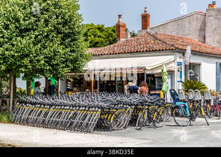 Ein Fahrradverleih mit dem Schild „Location de vélos“ steht auf dem Place Austerlitz in Le Bourg auf der Insel Aix (Île d'Aix), Charente-Maritime, Nouvelle-Aquitaine, Frankreich. Auf Englisch: „Fahrradverleih“. Lange Reihen von gelben und grauen Mieträdern stehen vor dem Gebäude mit seinem Ziegeldach, einige mit Kindersitzen ausgestattet. Kunden warten an der Theke unter dem Vordach. Die Insel ist autofrei und mit einer Länge von 3 Kilometern und einer Breite von 600 Metern perfekt für Radfahrer und Fußgänger. Stockfoto