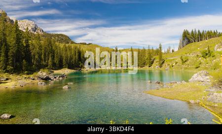 Das ruhige, klare türkisfarbene Wasser des Karersee, eingebettet zwischen dichten Kiefernwäldern und sanften grünen Hügeln, unter einem hellblauen Himmel mit hellem cl Stockfoto