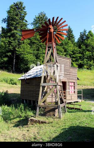 Eine hohe Windmühle aus Holz mit rostigen Metallklingen steht neben einer verwitterten Hütte mit rosa Vorhängen auf grasbewachsenem Boden und umgeben von dichtem Wasser Stockfoto