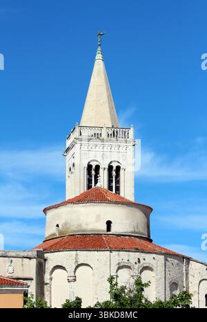 Ein hoher Kirchturm aus Stein mit spitzem Turm und Bogenfenstern erhebt sich über einem Rundschiff mit rot gekacheltem Dach vor einem klaren blauen Himmel in einem historischen Gebäude Stockfoto