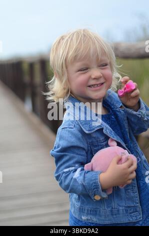 Das Kind lächelt fröhlich, während es ein rosafarbenes Spielzeugferkel auf einer Holzbrücke hält. Das Kind trägt eine Denim-Jacke und ein hellblaues Hemd, die Glück ausstrahlen Stockfoto