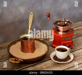 Tasse frisch gebrühten Kaffee auf einer Untertasse mit Bohnen, Handkaffeemühle und Tablett mit Sand- und Kupfercezve auf einem Holztisch. Draufsicht, flach. Stockfoto