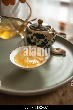 Hände, die Tee aus klarem Glaskrug in Teetassen gießen, schaffen ein ruhiges Teerritual, während sanftes Licht und hölzerner Teeteller an beruhigendes, achtsames Chinesisch erinnern Stockfoto