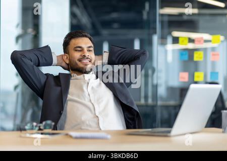 Ein lächelnder Geschäftsmann im Anzug entspannt sich in seinem Büro, lehnt sich mit den Händen hinter dem Kopf zurück und genießt seine Arbeit. Stockfoto