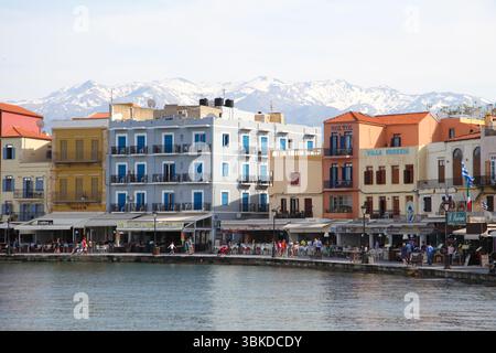 Die farbenfrohen Gebäude des alten Hafens von Chania mit Cafés und Marktständen am Ufer. Dahinter liegen die schneebedeckten Lefka Ori Berge Stockfoto