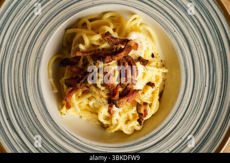 Eine wunderschön überzogene Portion Spaghetti Carbonara mit knusprigem Pancetta auf einer gestreiften Keramikplatte. Die cremige Soße und die goldenen Töne erzeugen einen Stockfoto
