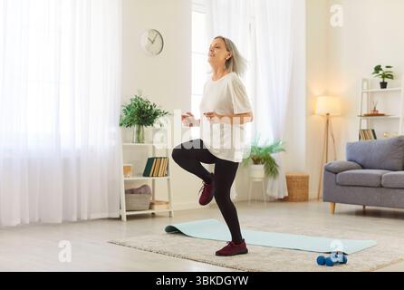 Lächelnde, reife Frau, die im Wohnzimmer vor Ort läuft, folgt dem Video-Tutorial für das Heimtraining. Stockfoto
