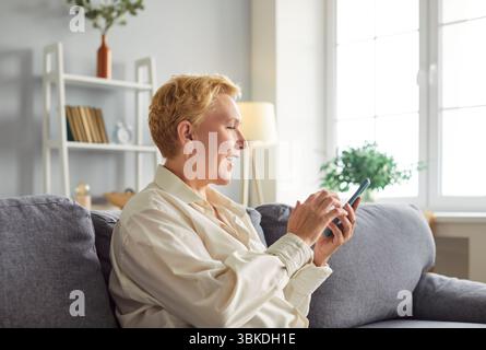 Fröhliche, reife Frau, die auf dem Sofa sitzt, soziale Medien scrollt und Online-Unterhaltung genießt. Stockfoto