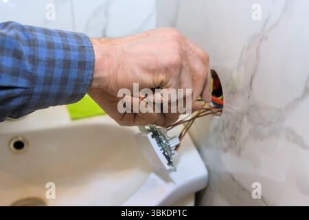 Der Auftragnehmer arbeitet an der Renovierung des Badezimmers und ersetzt den Lichtschalter, der an die freiliegende Verkabelung angeschlossen ist. Stockfoto