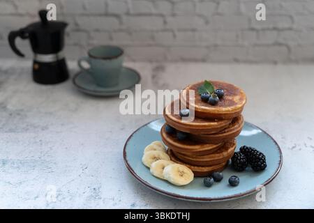 Leckerer Pfannkuchenstapel mit Beeren und Bananenscheiben. Eine Mokakanne und eine Kaffeetasse befinden sich in der Nähe auf der grauen Oberfläche und sorgen für eine warme Morgenstimmung Stockfoto