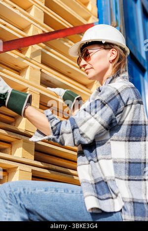 Porträt glückliche Fahrerin mit Sonnenbrille und Schutzhelm lächelt vor blauem Truck und gestapelten Paletten. Weibliche Person Mädchen Profi Stockfoto