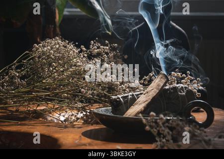 Palo Santo und Salbei verwischen mit sichtbarem Rauch, umgeben von getrockneten Blumen auf Holztisch Stockfoto