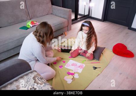Zwei Personen, ein Erwachsener und ein Kind, sitzen auf einer Fußmatte und kreieren Kunstwerke aus bunten Papierherzen. Sie sind an einem Handwerksprojekt beteiligt und fördern Stockfoto