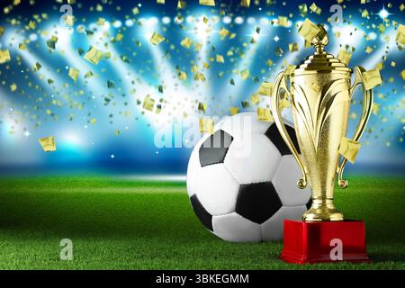 Fußballmeisterschaft. Glänzend goldener Trophäenbecher und Fußball unter fallendem Konfetti im Stadion Stockfoto