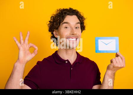 Junger Mann, der ein Postsymbol auf gelbem Hintergrund zeigt, während er lächelt und mit der Hand ein OK-Zeichen drückt Stockfoto