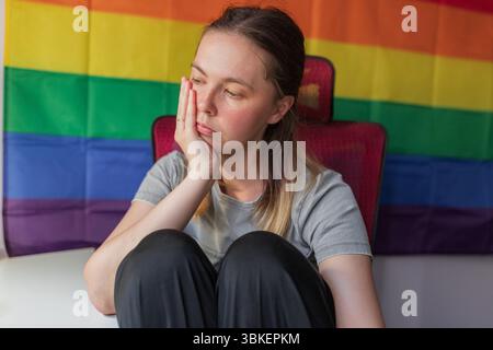 Eine nachdenkliche Frau, die Kopf auf der Hand liegt, vor dem Hintergrund des Regenbogenstolzes sitzt. Die Frau sitzt mit gezogenen Knien, das Gesicht auf der Hand liegend, nachdenkliche Expensionen Stockfoto