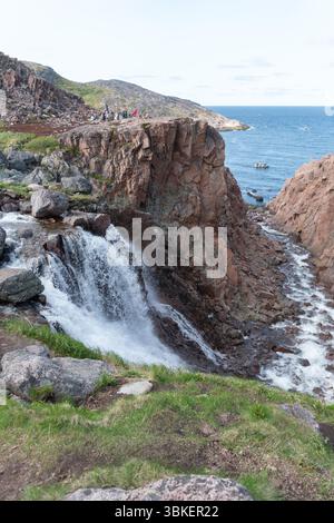Teriberka, Russland - 13. Juni 2025: Blick auf den Battery Wasserfall, der durch raue rote Felsen in die Barentssee stürzt, mit kleinen Touristengruppen Stockfoto