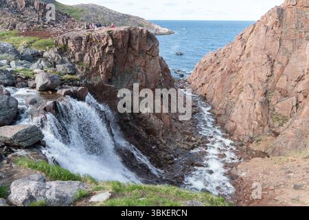 Teriberka, Russland - 13. Juni 2025: Blick auf den Battery Wasserfall, der durch raue rote Felsen in die Barentssee stürzt, mit kleinen Touristengruppen Stockfoto