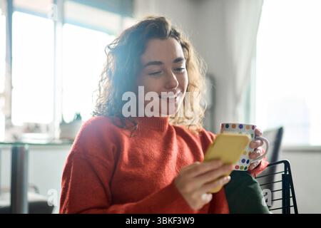 Junge glückliche Frau, die ihr Handy benutzt und Kaffee trinkt. Stockfoto