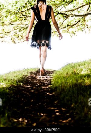 Eine Frau in einem schwarzen Kleid läuft barfuß auf einem Weg durch ein grasbewachsenes Gebiet. Stockfoto