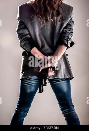 Eine Person, von hinten gesehen, hält eine Waffe hinter ihrem Rücken. Sie tragen einen Blazer und Jeans. Stockfoto