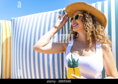 Eine junge Frau, die ihren Strohhut verstellt, während sie einen tropischen Cocktail in einem ananasförmigen Glas in einem Strandresort hält Stockfoto