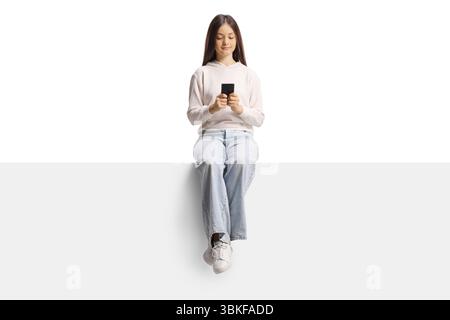 Weiblicher Teenager, der auf einem leeren Panel sitzt und ein Smartphone auf weißem Hintergrund benutzt Stockfoto