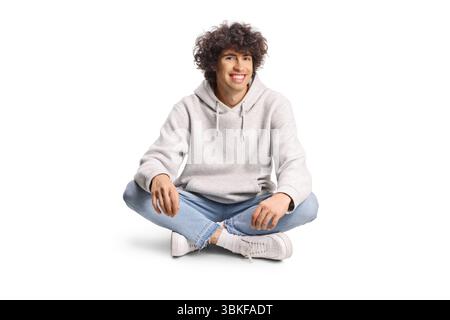 Junger Mann mit lockigen Haaren, der mit überkreuzten Beinen auf weißem Hintergrund sitzt Stockfoto