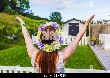 Mädchen mit Blumenkranz hebt die Arme während der schwedischen Mittsommerfeier freudig im Garten. Schweden. Stockfoto