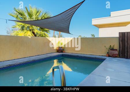 Swimmingpool im Hinterhof mit Dreieck-Sonnenschirm segeln darüber. Gelbe Mauer und Palmen im Hintergrund. Stockfoto