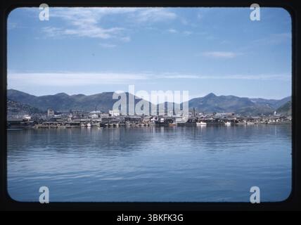 Blick auf die Uferpromenade von Masan, Südkorea, aufgenommen im Oktober 1956. Traditionelle koreanische Hafengebäude und kleine Boote säumen die Küste, die von den sanften Hügeln und Gebirgszügen der südkoreanischen Halbinsel flankiert werden. Dieses Bild zeigt eine seltene Farbaufnahme der Hafeninfrastruktur Masans während der Zeit der Erholung nach dem Koreakrieg. Stockfoto