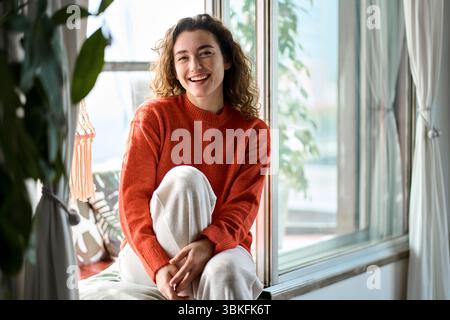 Glückliche, hübsche junge Frau, die zu Hause auf dem Fensterbrett sitzt und lacht. Stockfoto