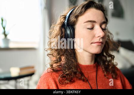 Eine junge, aufmerksame Frau mit Kopfhörern, die zu Hause Meditation macht. Stockfoto