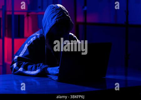 Ein Mann in einem schwarzen Kapuzenpullover, der in einem neonbeleuchteten Raum einen Laptop benutzt und sein Gesicht im Schatten versteckt hat. Er erinnert an Themen wie Cybersicherheit, Hacking, Codin Stockfoto