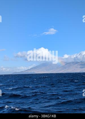 Ein schwacher Regenbogen erscheint über den Bergen von Maui, vom Meer aus gesehen an einem klaren Tag. Stockfoto