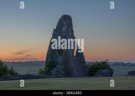 Benfleet, Großbritannien. Juni 2025. Die Überreste einer teilweise eingestürzten Steinstruktur stehen während des Sonnenaufgangs auf einem grasbewachsenen Feld. In der erhaltenen Mauer sind zwei Fensteröffnungen sichtbar. Die Szene erinnert an Themen wie Geschichte, Verfall und natürliche Schönheit, während warmes Licht lange Schatten über die Landschaft wirft. Die Menschen versammeln sich in der Ruine von Hadleigh Castle, um den Sonnenaufgang bei der Sommersonnenwende zu beobachten. Penelope Barritt/Alamy Live News Stockfoto