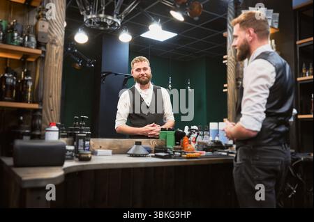 Erfahrener Friseur bereitet den Platz für den nächsten Kunden vor. Friseurreinigungstabelle nach dem letzten Kunden. Hübscher Mann, der in den Spiegel blickt und lächelt. Stockfoto