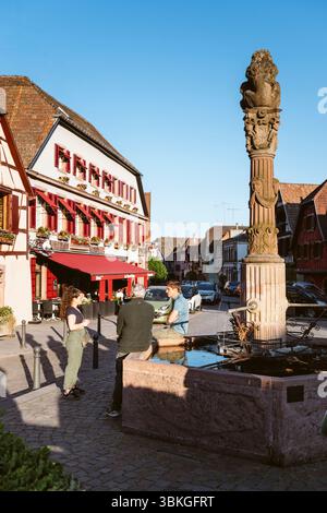 Jugendliche unterhalten sich in der Abendsonne am Brunnen auf dem Platz der Republik vor den Fachwerkhäusern von Ribeauvillé, Elsass, Frankreich Stockfoto