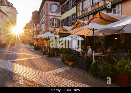 Ribeauvillé, Elsass, Frankreich: Gäste auf der Terrasse der Brasserie La Post in der mittelalterlichen Gasse mit Fachwerkhäusern, die vom Sonnenuntergang beleuchtet werden Stockfoto